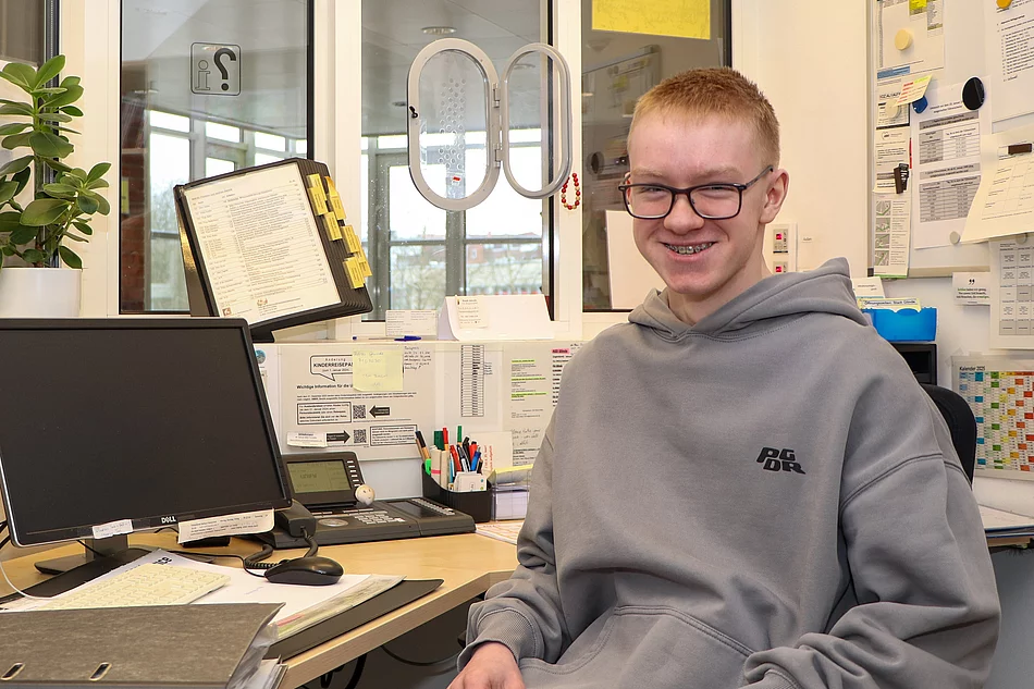 Ein Jugendlicher mit Brille sitzt an einem Schreibtisch und lächelt.