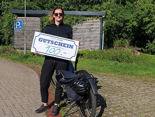 Ulrike Wegner vom Team FEINSTAUBVERMEIDER Eine Frau halt einen sehr großen Gutschein mit der Zahl 100 in Händen und steht an einem Fahrrad.