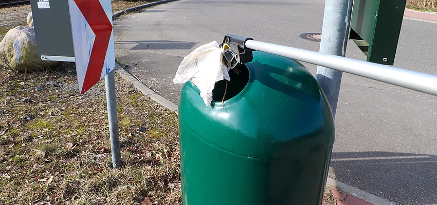 Ein grüner Mülleimer in den mit eine Zange Müll geworfen wird.