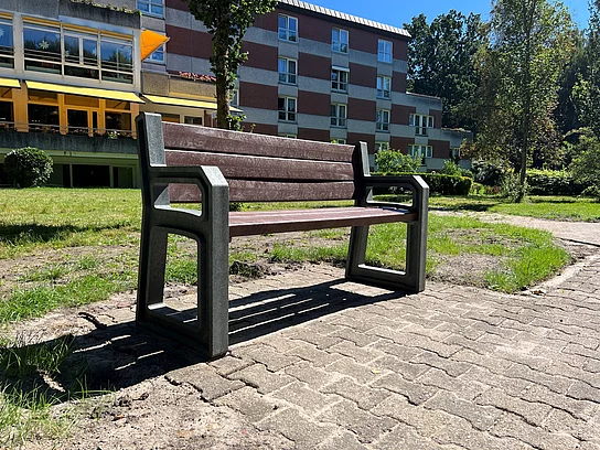 Eine erhöhte Parkbank mit Armlehnen. Im Hintergrund das Pflegeheim Haus Togohof