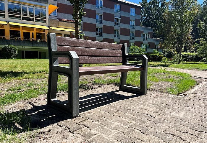 Eine erhöhte Parkbank mit Armlehnen. Im Hintergrund das Pflegeheim Haus Togohof