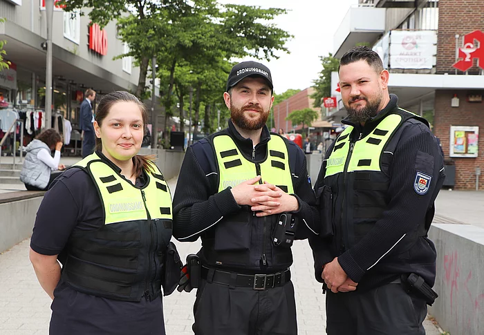 Eine Frau und zwei Männer in blauen Uniformen mit einer reflektierenden Warnweste stehen nebeneinander. 