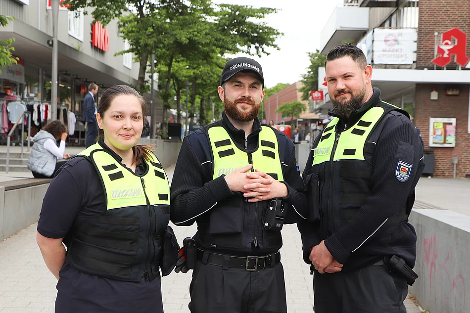 Team des Kommunalen Ordnungsdienstes Glinde Eine Frau und zwei Männer in blauen Uniformen mit einer reflektierenden Warnweste stehen nebeneinander.