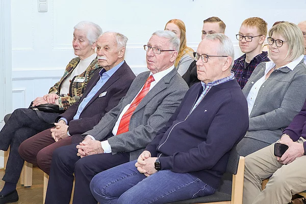 Festlich gekleidete Menschen sitzen nebeneinander auf Stühlen und schauen nach vorn.