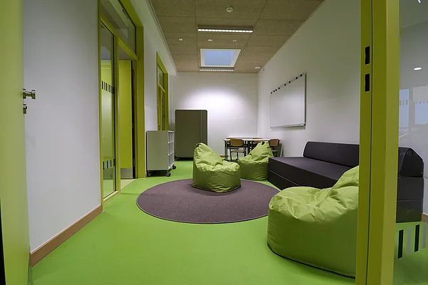 Immer zwei Klassenräumen teilen sich einen Differenzierungsraum mit Sitzsäcken, Sofa und Arbeitsplätzen. Der Raum ist in einem frischen Grün gehalten und hat ein Oberlicht und neben den Türen Fenstern.