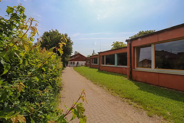 links grüne Büsche, rechts ein rotes flaches Gebäude mit großen Fenstern, in dem der Hort Löwenzahn Tannenweg untergebracht ist.
