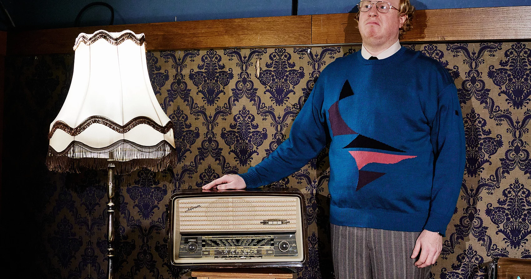 Ein Mann mit blauem Pullover steht neben einem alten Radio und einer Stehlampe.