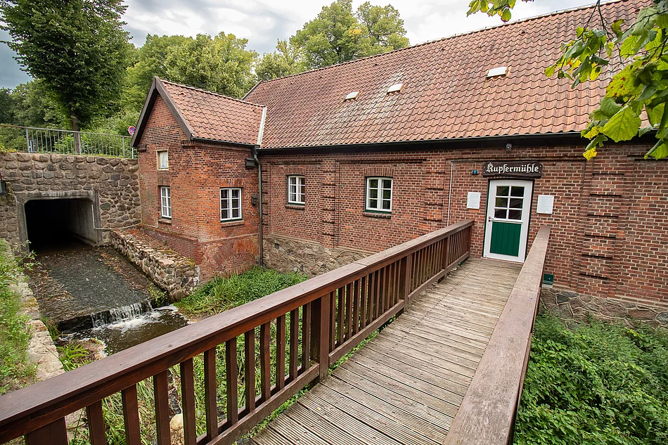 Kupfermühle Glinde (2024) Ein Backsteingebäude mit Satteldach (Kupfermühle) mit Holzbrücke über die Glinder Au.