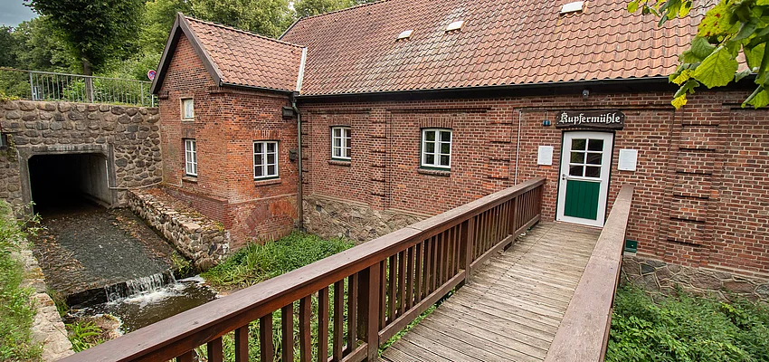 Ein Backsteingebäude mit Satteldach (Kupfermühle) mit Holzbrücke über die Glinder Au.