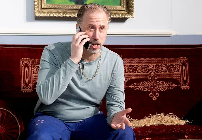 Ein Mann mit Arbeitshose sitz auf einer Couch und telefoniert überrascht.