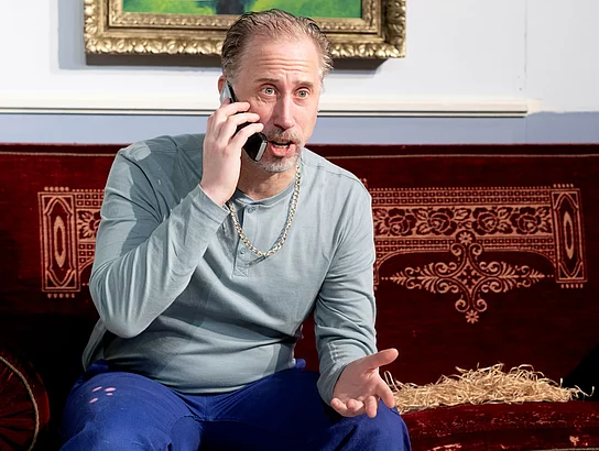 Ein Mann mit Arbeitshose sitz auf einer Couch und telefoniert überrascht.
