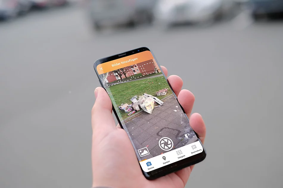 Eine Hand hält ein Handy in der Hand, darauf ein Foto mit Müll.