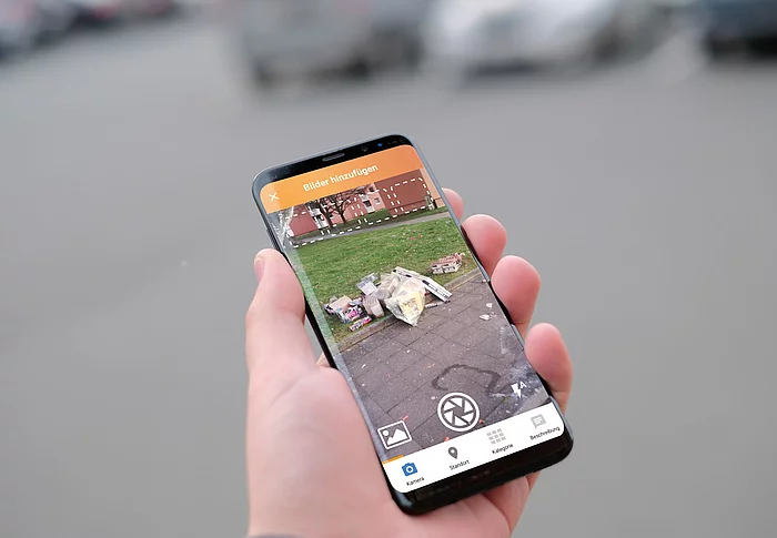 Eine Hand hält ein Handy in der Hand, darauf ein Foto mit Müll.