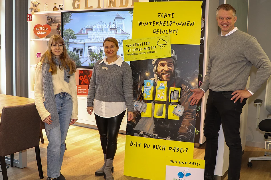 Zwei Frauen und ein Mann stehen vor einem gelben Aufsteller mit Werbekarten.