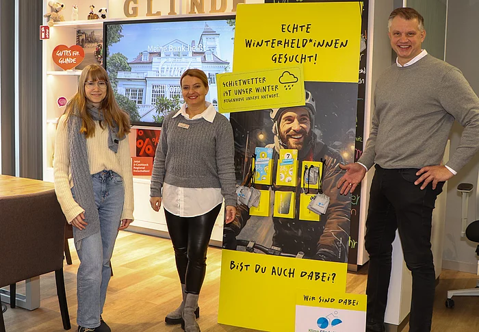 Zwei Frauen und ein Mann stehen vor einem gelben Aufsteller mit Werbekarten.
