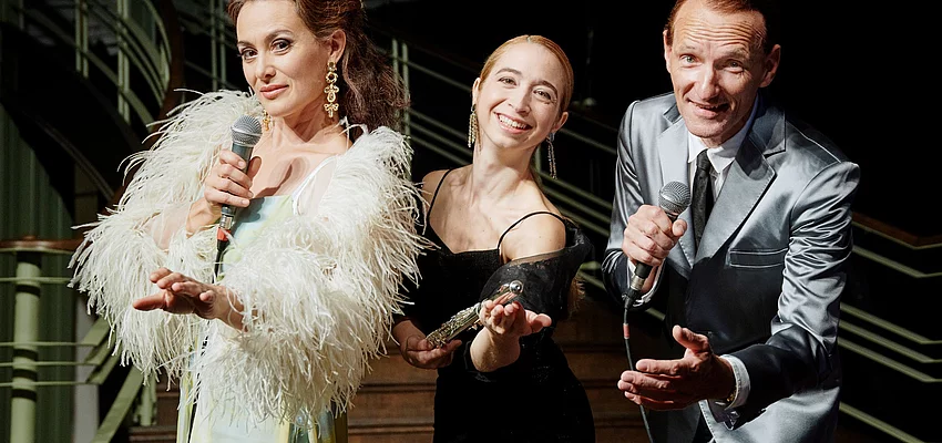 Zwei Frauen und ein Mann im Gala-Outfit mit Mikrofonen in der Hand.