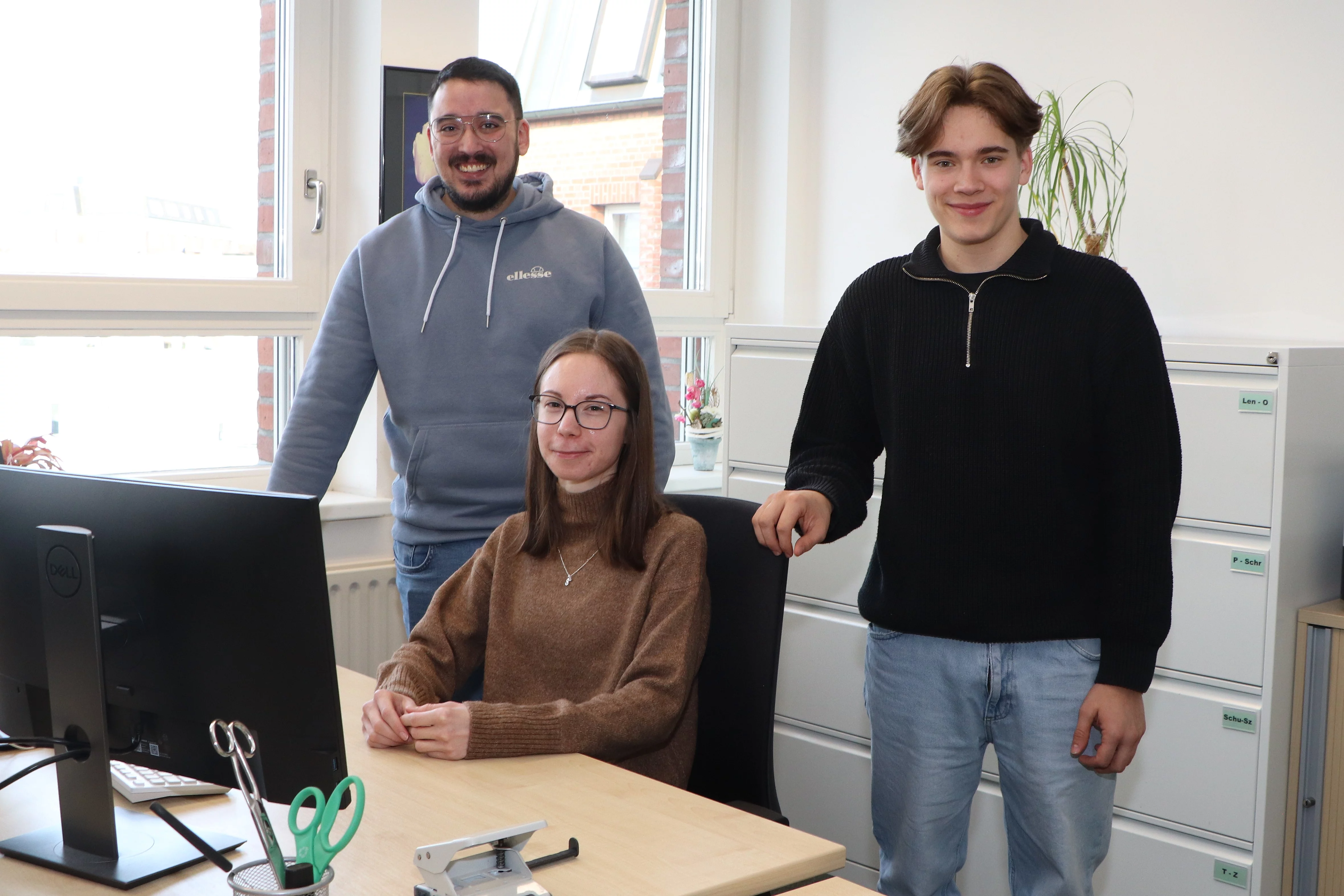 An einem Schreibtisch sitzt eine junge Frau, hinter ihr stehen zwei junge Männer.