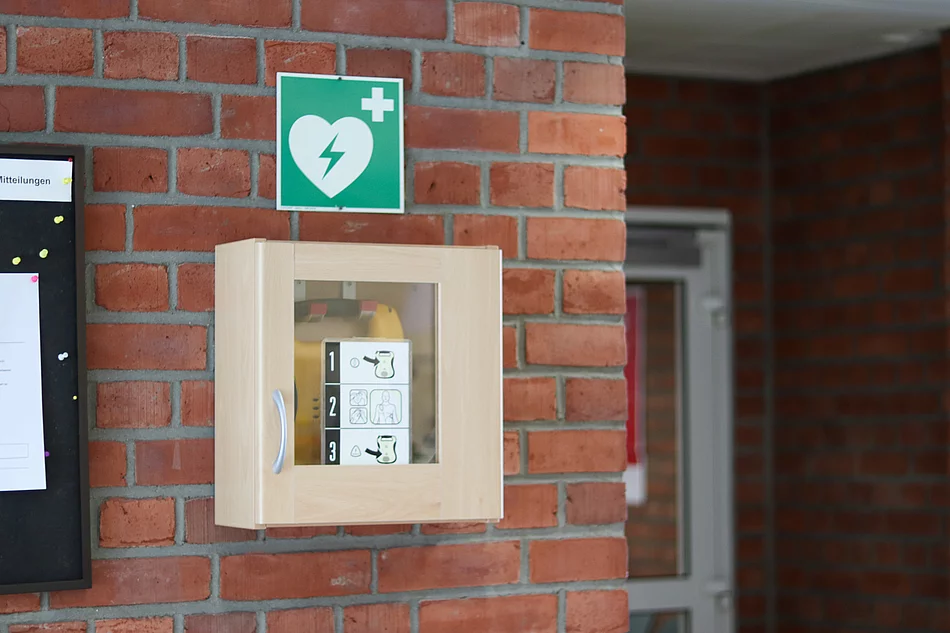 Defibrillator im Eingangsbereich des Rathauses In einem kleinen Schrank ist ein gelber Defibrillator, darüber ein grünes Schild mit weißem Herz und Blitz.