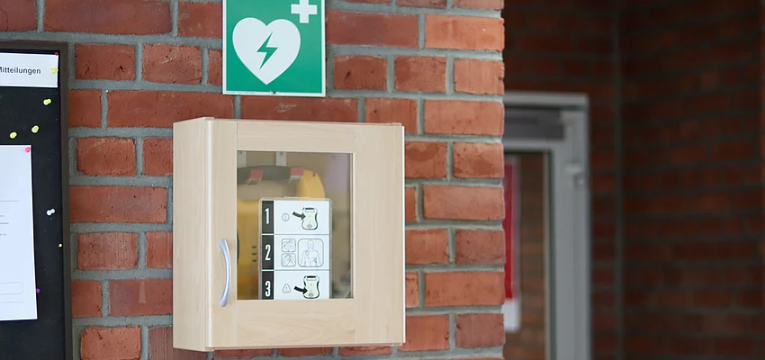 Defibrillator im Eingangsbereich des Rathauses In einem kleinen Schrank ist ein gelber Defibrillator, darüber ein grünes Schild mit weißem Herz und Blitz.