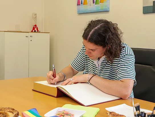 Eine junge Frau mit lockigen braunen Haaren beugt sich über ein Buch und schreibt etwas.