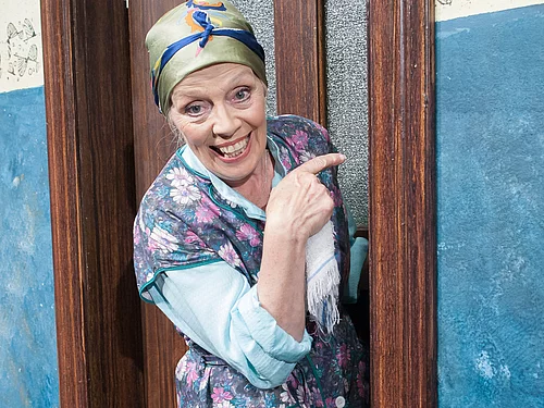 Eine ältere Frau mit Kopftuch und Kittel schaut lächelnd aus einer Tür.