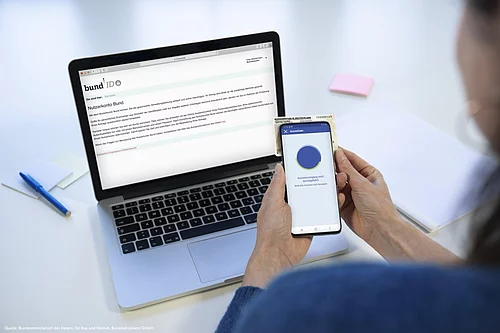 Auf einem Bildschirm ist die Seite BUND ID zu sehen. Davor ein Mobiltelefon mit geöffneter APP. Das das Telefon wird ein Ausweis gehalten. 