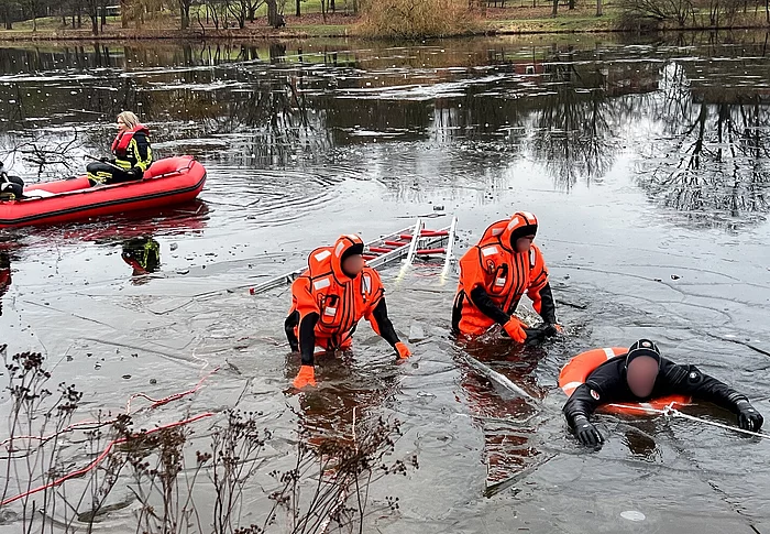 Eine Person mit Taucheranzug wird von zwei weiteren Personen im Taucheranzug aus einem See mit Eisschollen gerettet.