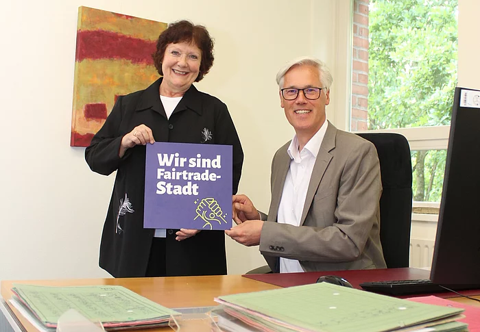 Ulrike Müller Kopsch (Vorsitzende der Fairtrade Steuerungsgruppe Glinde) und Bürgermeister Rainhard Zug an einem Schreibtisch mit einem Schild "Wir sind Fairtrade-Stadt".