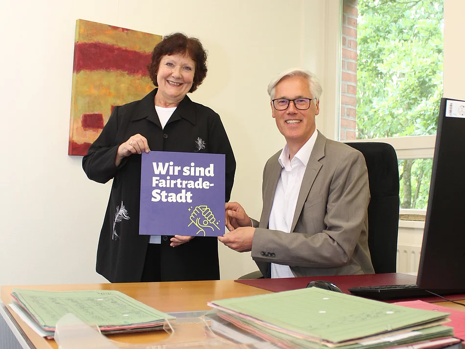 Ulrike Müller Kopsch (Vorsitzende der Fairtrade Steuerungsgruppe Glinde) und Bürgermeister Rainhard Zug an einem Schreibtisch mit einem Schild "Wir sind Fairtrade-Stadt".