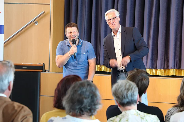 Zwei lächelnde Männer vor Publikum.