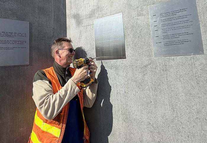 Ein Mann mit orangefarbener Weste und Akkuschrauber befestigt ein Schild an einer Betonwand.