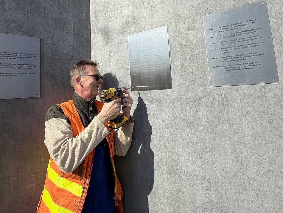 Ein Mann mit orangefarbener Weste und Akkuschrauber befestigt ein Schild an einer Betonwand.
